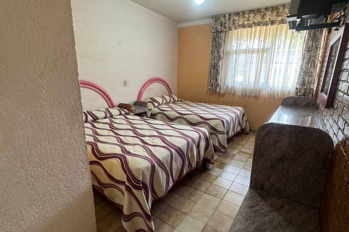 Ein Zimmer mit zwei Betten, einem Deckenventilator, einem Fernseher und einem Fenster mit Vorhängen.