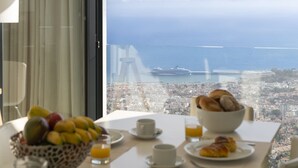 Une table de petit-déjeuner avec des fruits, des croissants et du jus d’orange, donnant sur un paysage urbain et la mer.