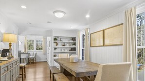 Un comedor con una mesa de madera, bancos y sillas. Hay una ventana con cortinas, un estante con objetos decorativos y una lámpara a la izquierda.