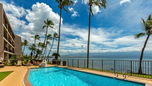 A swimming pool with a view of the ocean, surrounded by palm trees and a building with balconies. - Oceanfront Maui condo with lanai, pool and parking summer deals (Wailuku)