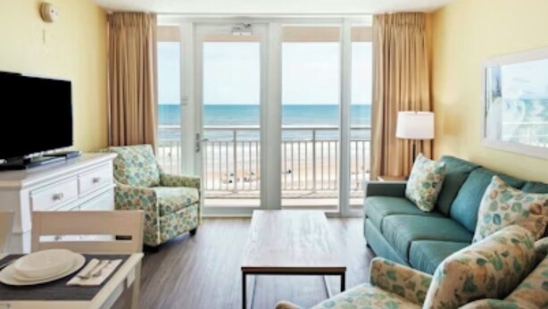 A room with a sofa, a television, a dining table with chairs, and a view of the beach through large glass doors. - Luxury Coastal Escape Beachfront Daytona Bike Week (Ormond Beach)