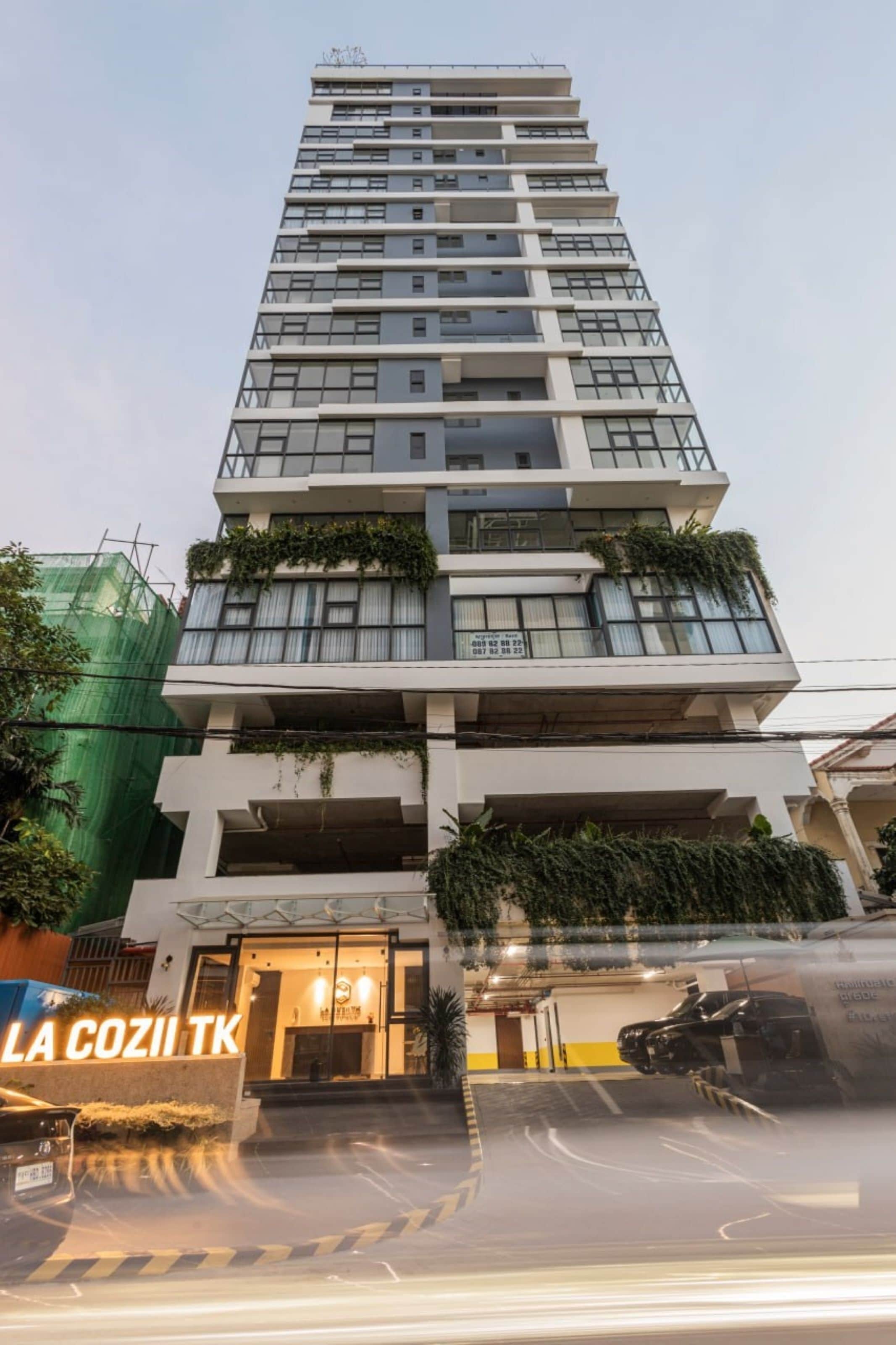 A multi-story building with a modern design, featuring balconies with greenery. The entrance has a sign and a parking area is visible in front.