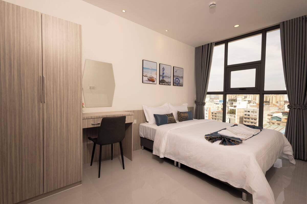 A modern hotel room with a bed, desk, and chair, large windows with curtains, and artwork on the wall.