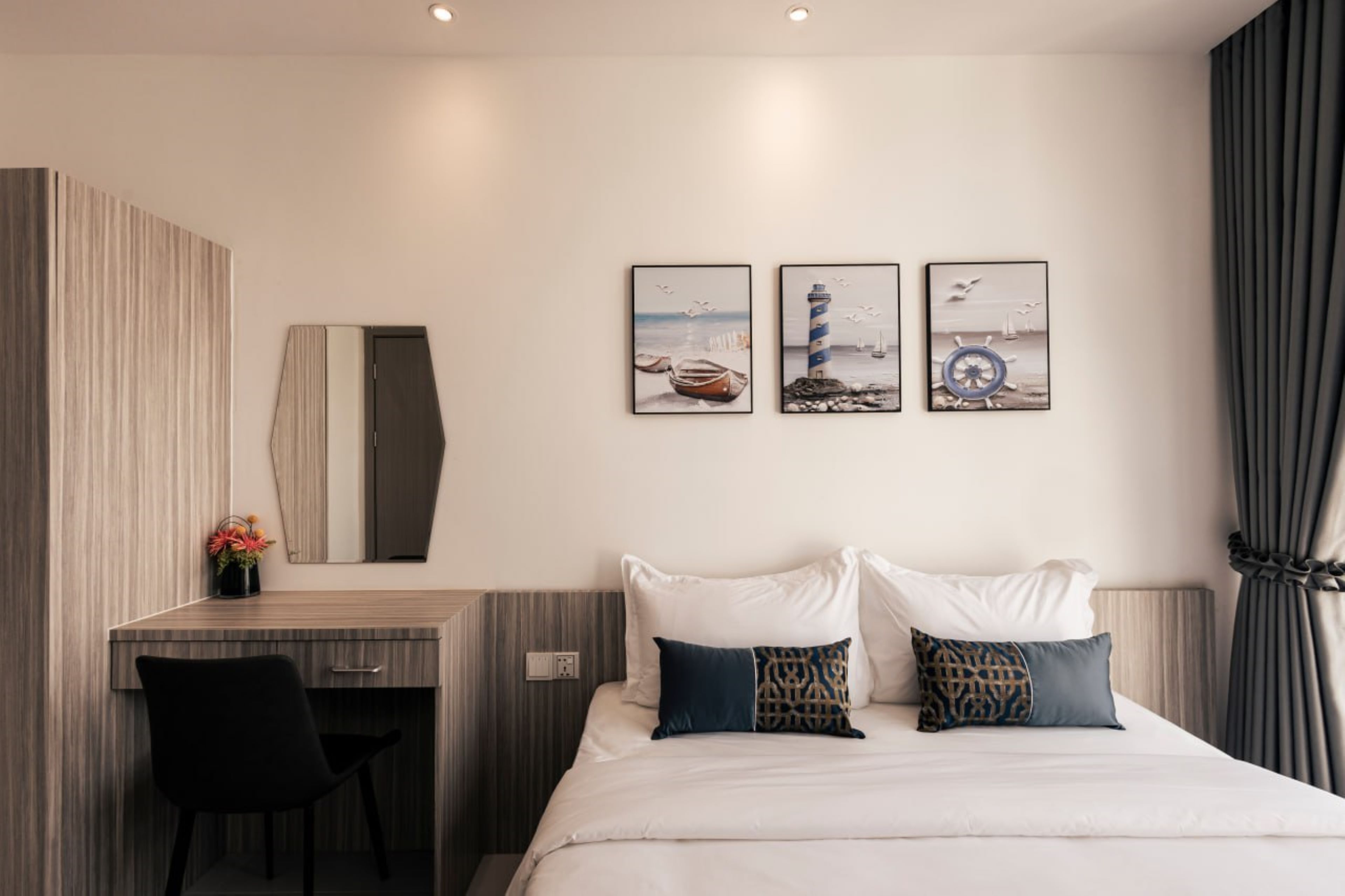 A modern hotel room with a bed, a desk, and a large mirror. There are three framed pictures on the wall above the bed.