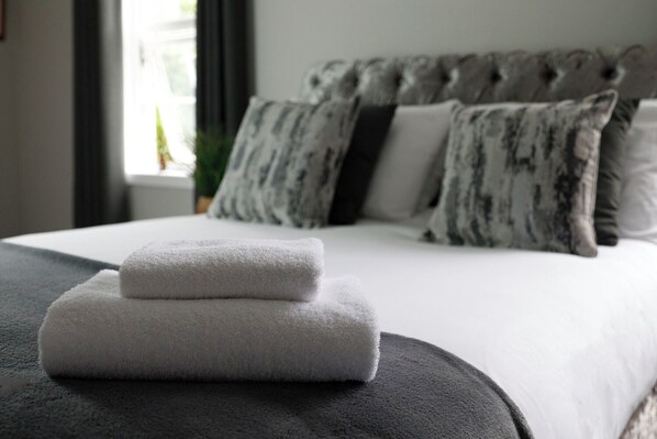 A neatly made bed with white linens and two folded towels on a dark ottoman.