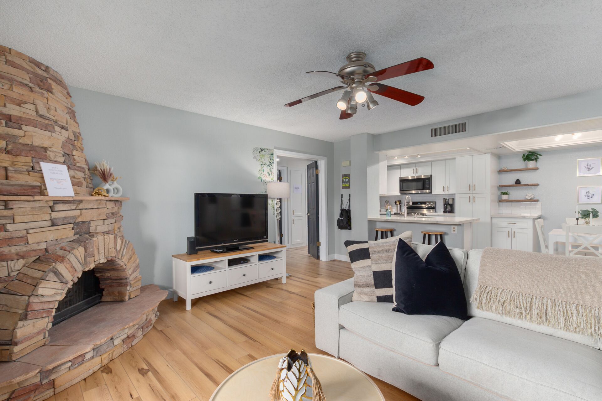 A living room with a stone fireplace, a flat-screen TV, a ceiling fan, and a light-colored sofa.