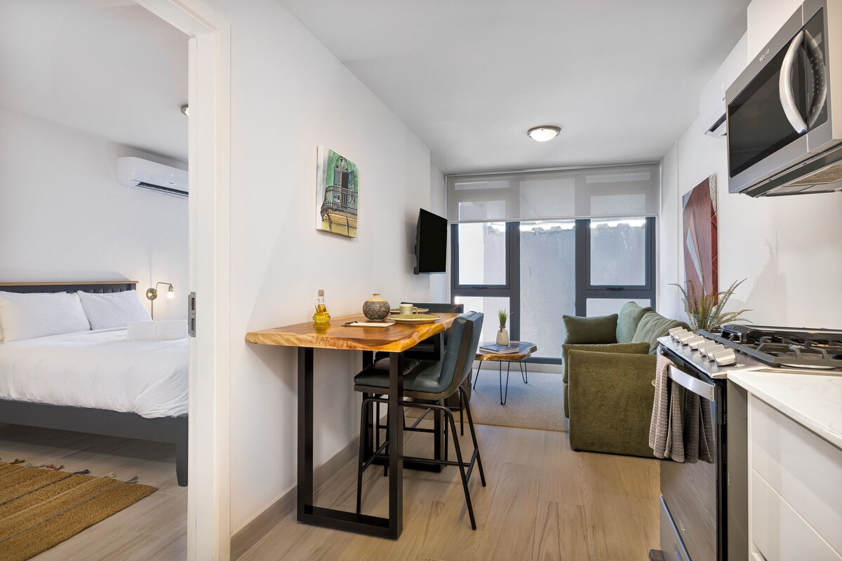 A compact hotel room with a bed, a dining area with a table and chairs, a kitchenette, and a large window providing natural light.