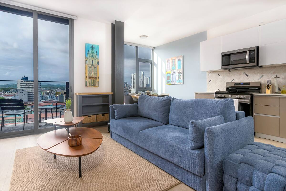 A modern living room with a sofa, coffee table, and a view of a cityscape through large windows.