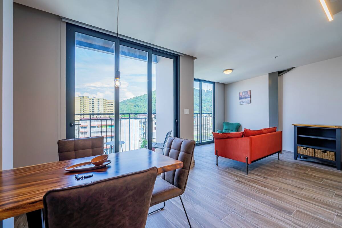 A modern living space with a dining area, a sofa, and a view of buildings through large windows.