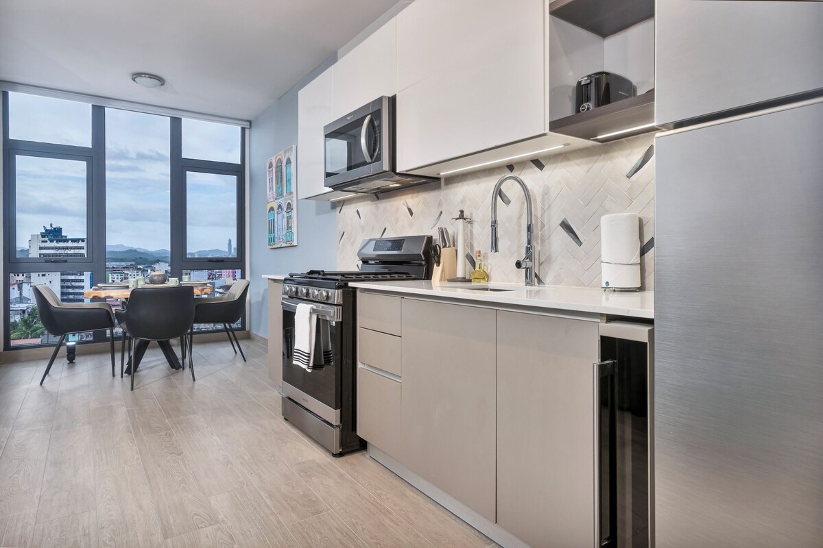 A modern kitchen with a large window, stainless steel appliances, and a dining area with a view.