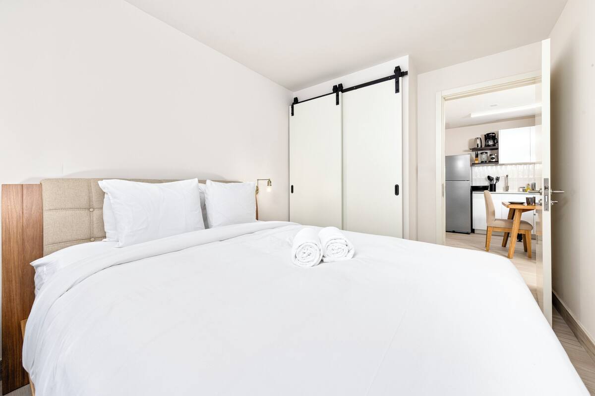 A hotel room with a large bed, a wooden headboard, and a kitchenette in the background.
