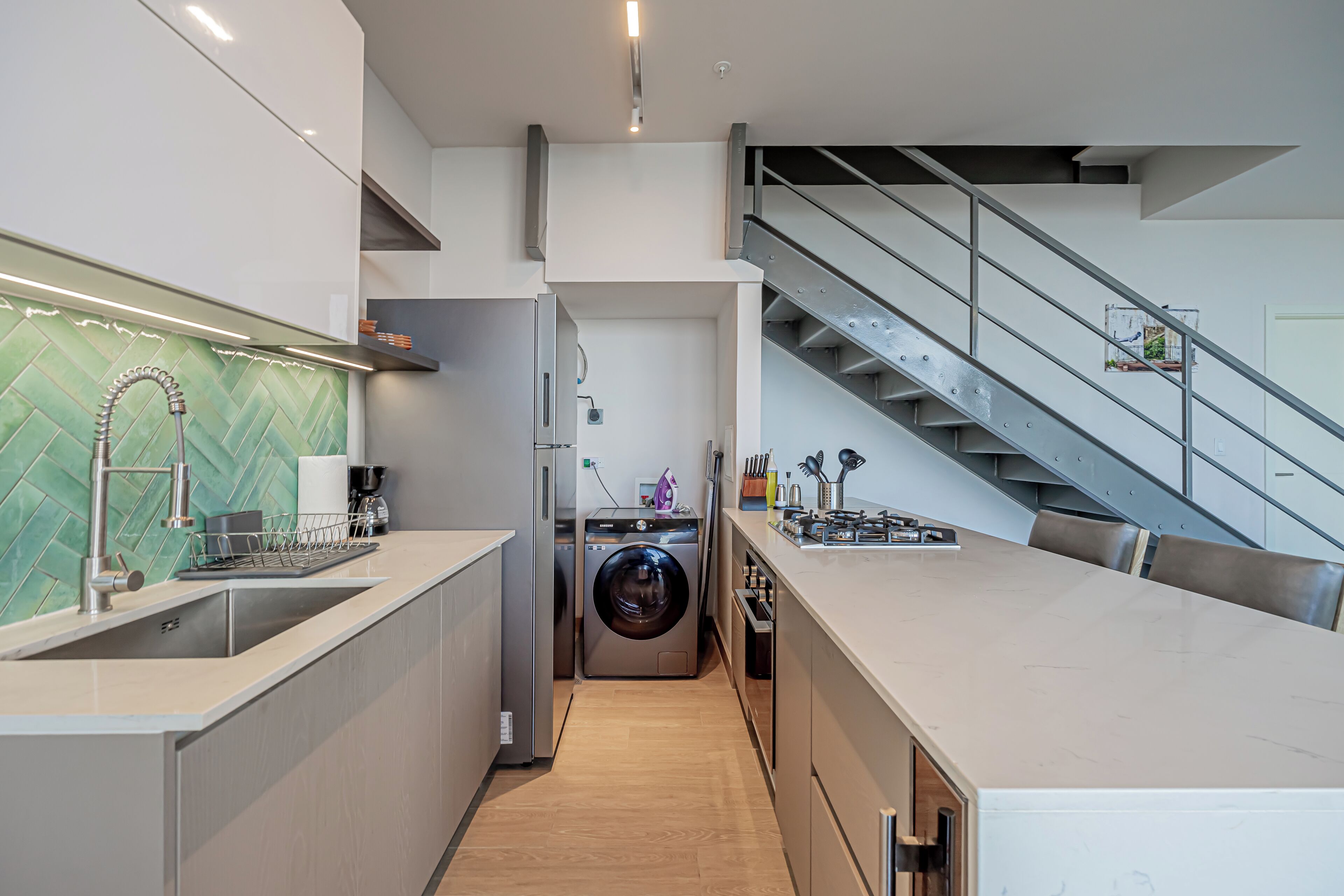 A modern kitchen with a built-in washer and dryer, a stainless steel sink, and a staircase leading to an upper level.