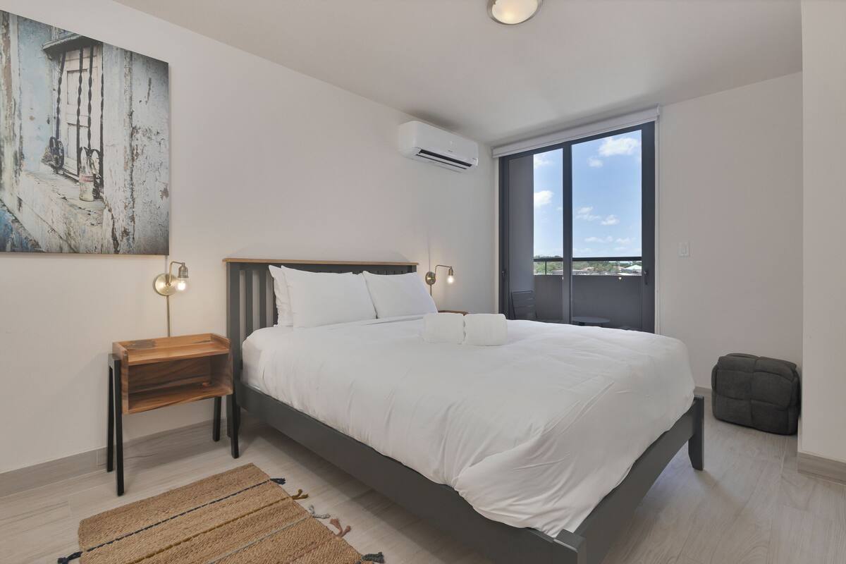 A modern bedroom with a large bed, a small bedside table, a wall-mounted air conditioner, and a large window offering a view.
