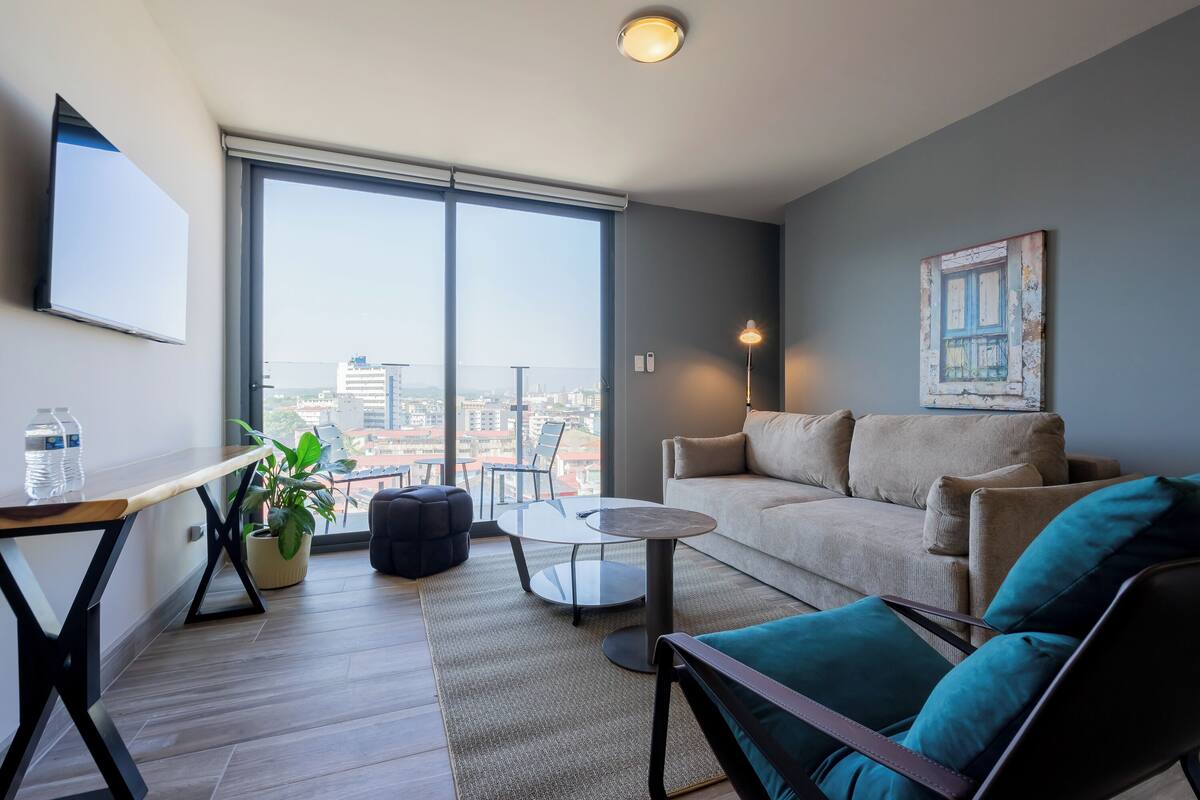 A modern living room with a large window, a sofa, a coffee table, and a TV mounted on the wall.