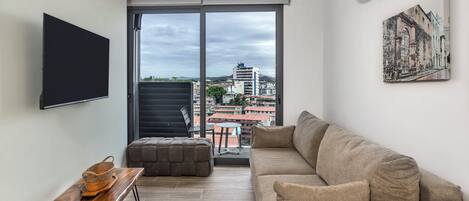 Una sala de estar moderna con sofá, televisión de pantalla plana, mesa de madera y balcón con vistas a la ciudad.