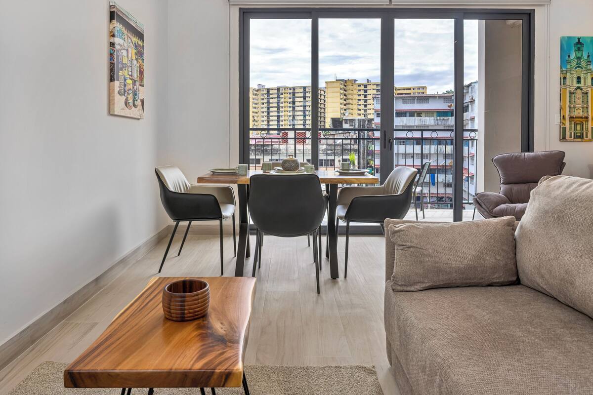 A modern living room with a dining area, large windows, and a view of high-rise buildings.