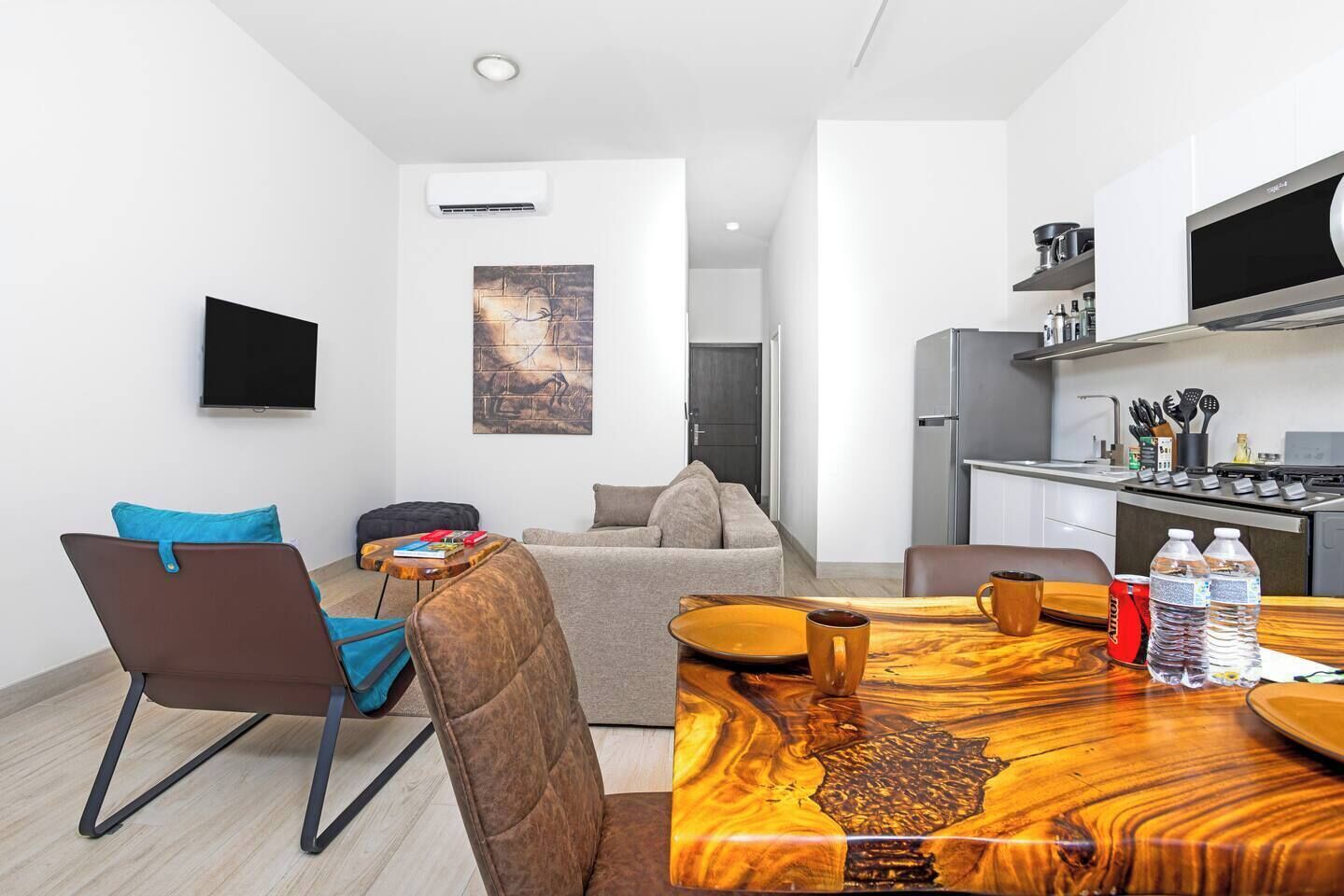 A modern living room with a sofa, a coffee table, and a dining table with chairs. There is a kitchen area with a refrigerator, stove, and microwave.