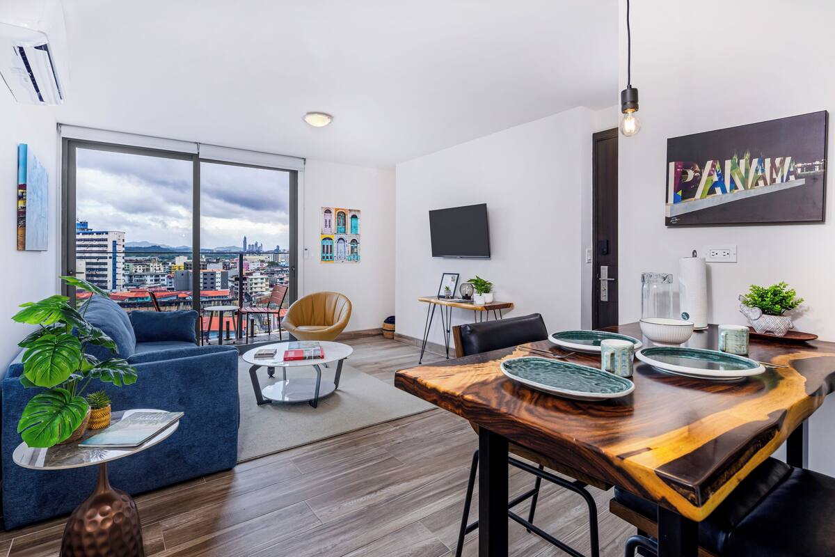 A modern living room with a large window, a dining area with a wooden table, and a city view.