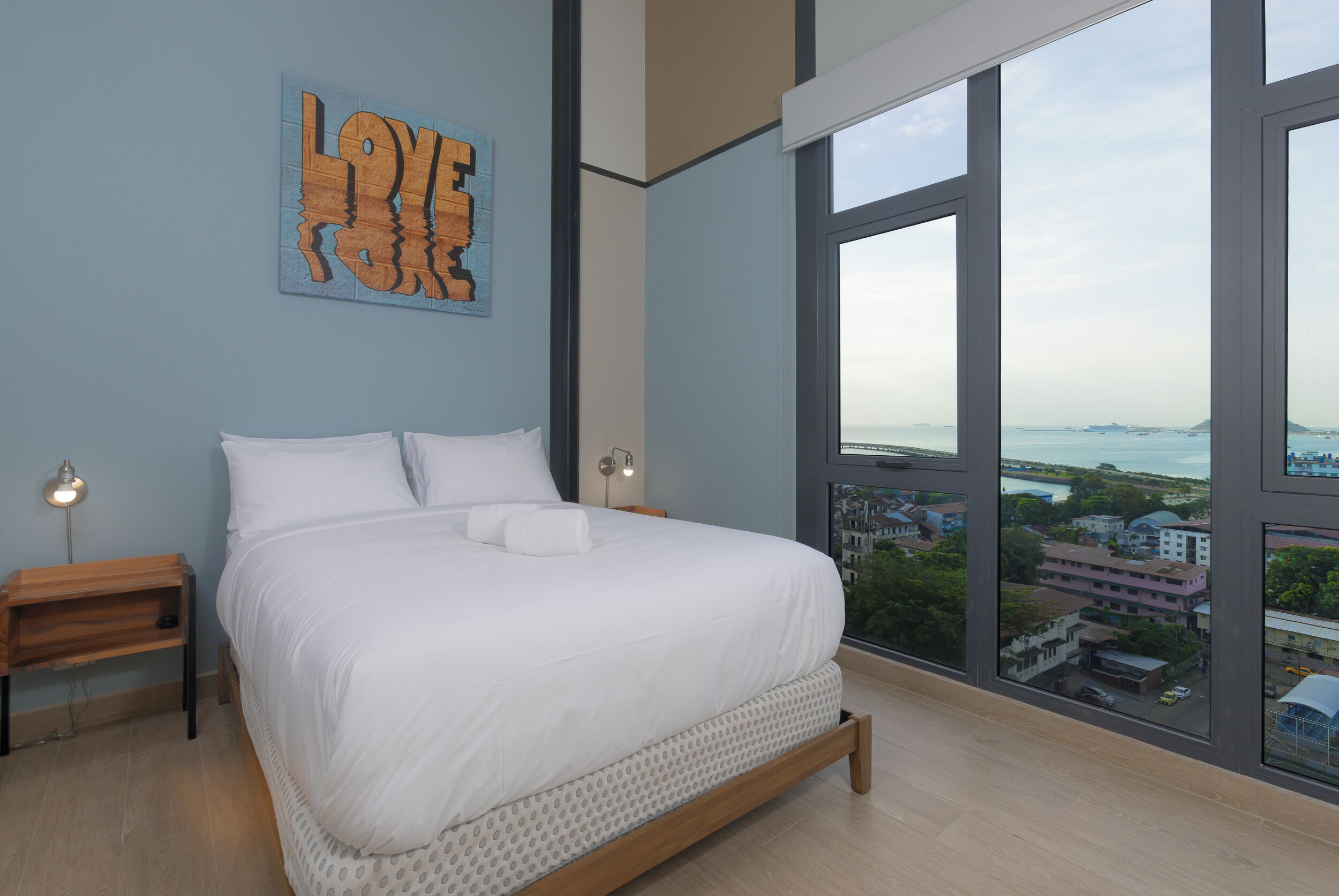 A bedroom with a large window, a bed, a nightstand, and a wall art piece.