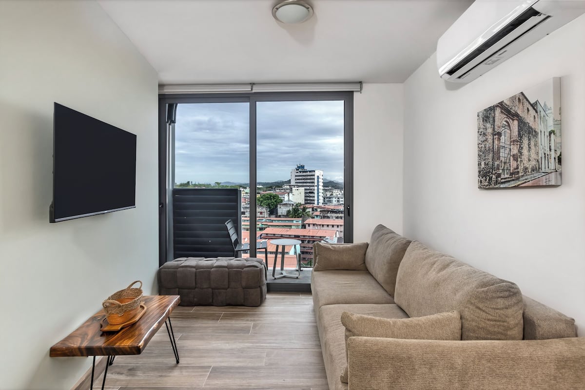 A modern living room with a sofa, a flat-screen TV, a wooden table, and a balcony with a city view.