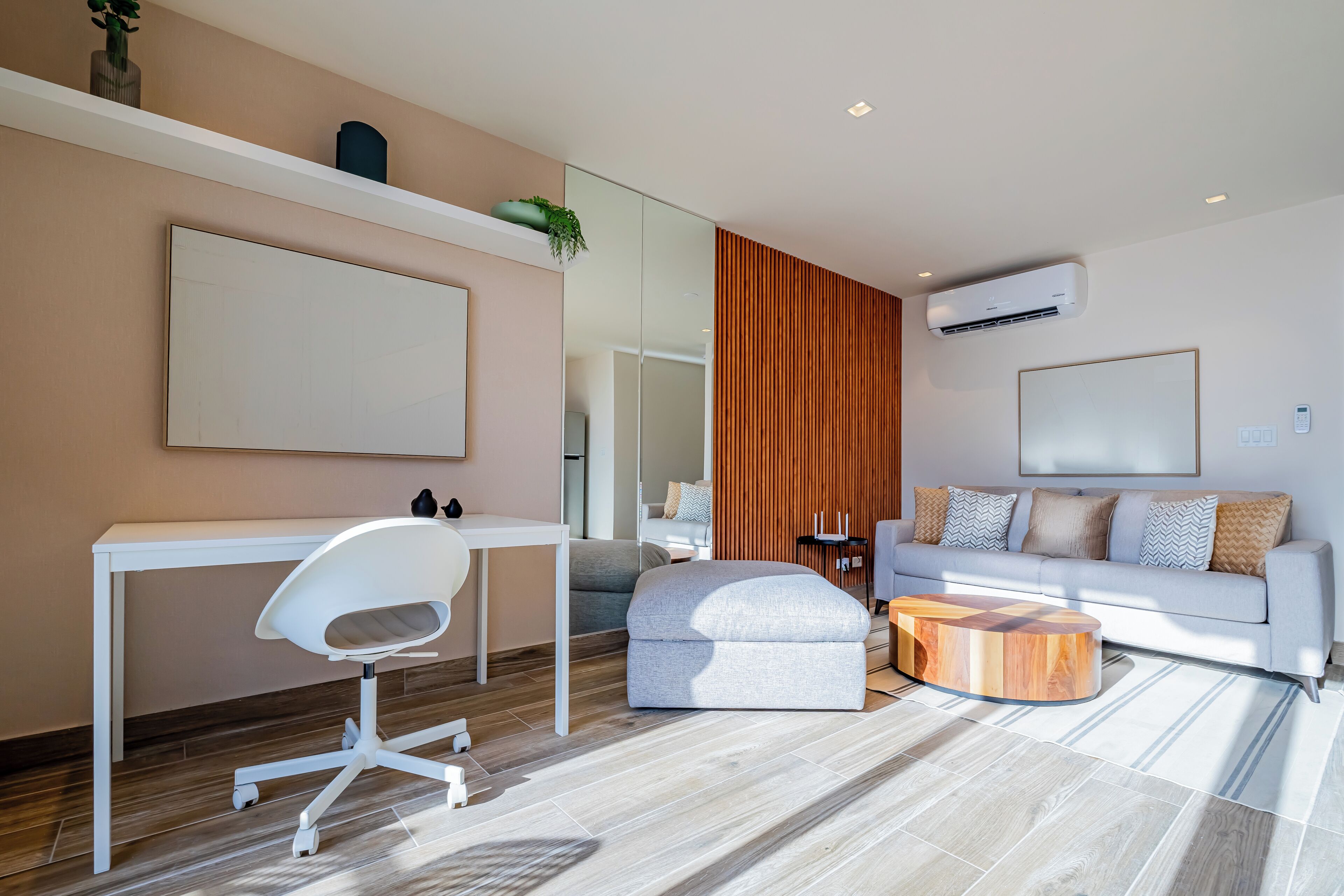 Una sala de estar moderna con un sofá blanco, una mesa de centro de madera, un escritorio blanco con una silla y un espejo grande en la pared.