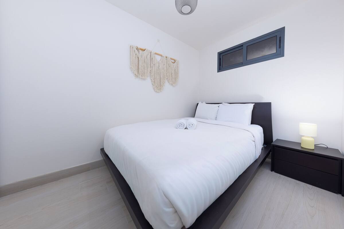 A neatly made bed with white linens, a bedside table with a lamp, and a macrame wall hanging.