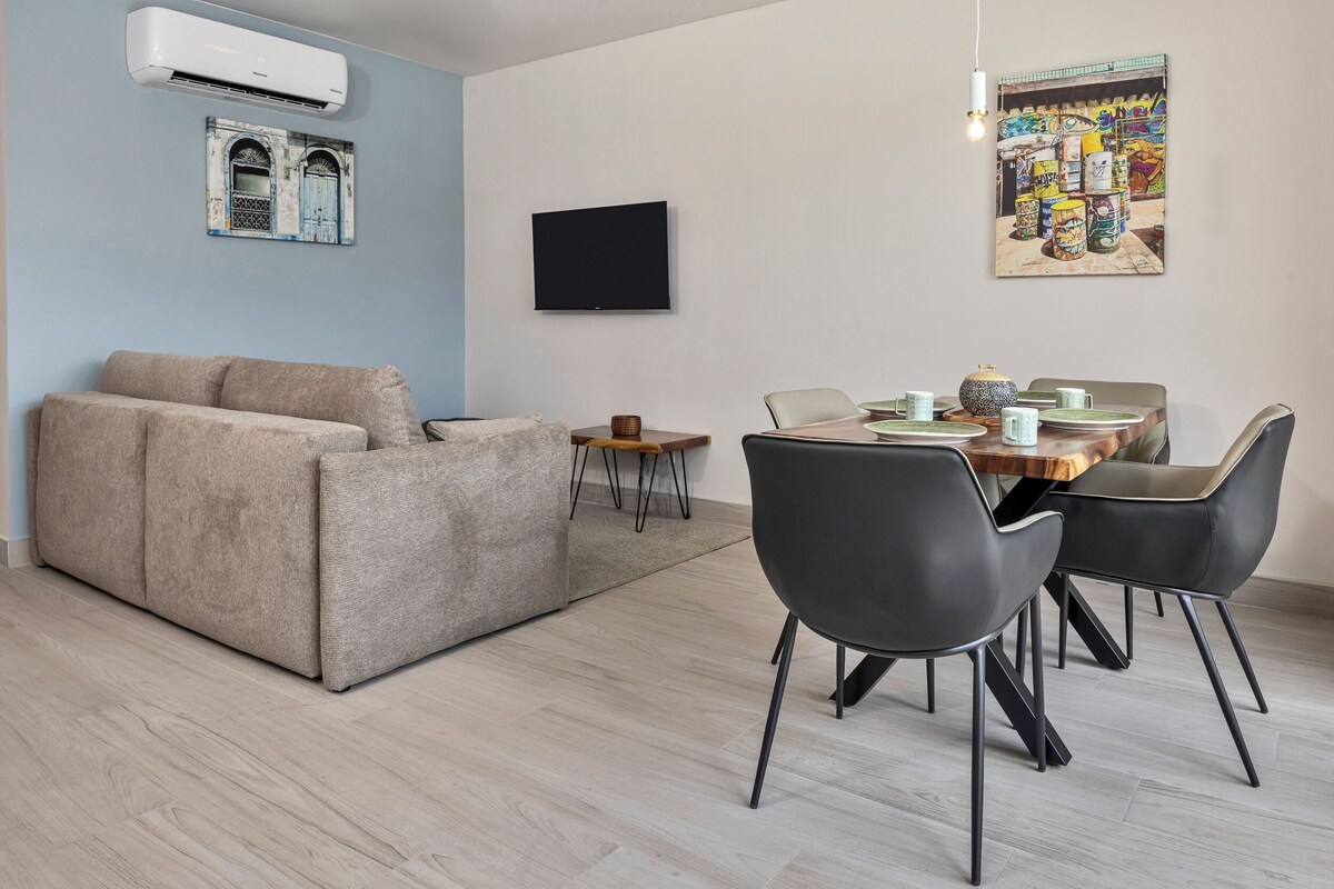 A modern living room with a beige sofa, a dining table with chairs, a flat-screen TV, and a wall-mounted air conditioner.