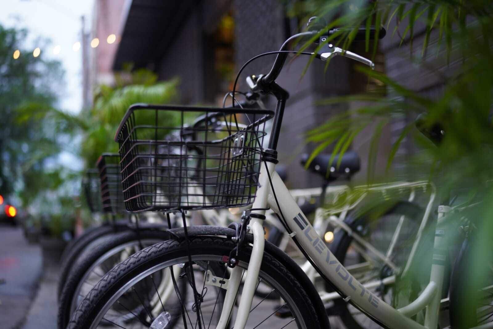 Un vélo avec un panier à l’avant, garé sur un trottoir, avec de la verdure et des bâtiments en arrière-plan.
