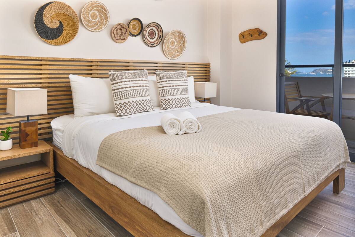 A neatly made bed with a beige blanket, two pillows with patterned designs, and rolled towels. There are woven wall decorations above the bed and a wooden headboard.