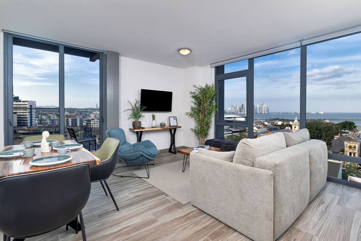 A modern living room with a dining area, large windows, a flat-screen TV, and a city view.