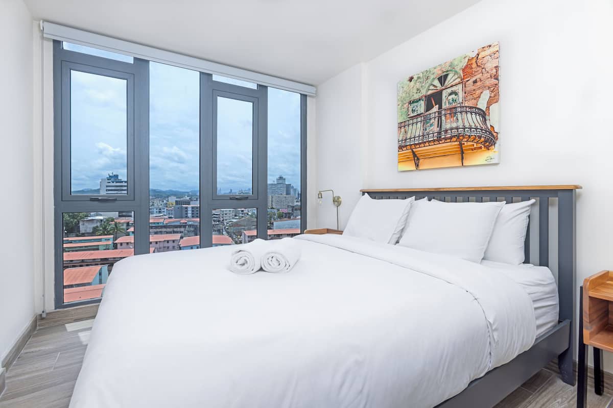 A hotel room with a large bed, white bedding, a city view through floor-to-ceiling windows, and a painting on the wall.