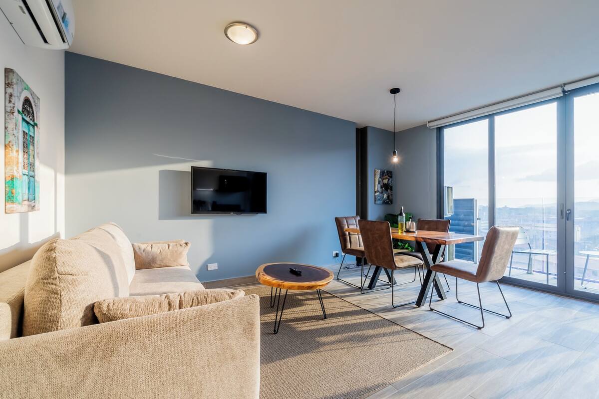 A modern living room with a sofa, a dining table with chairs, a TV mounted on the wall, and a large window offering a city view.