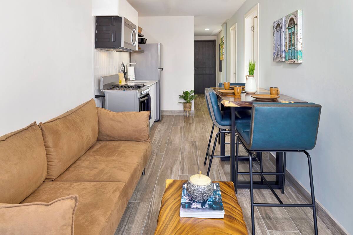 A modern living room with a brown sofa, a wooden coffee table with a book and a decorative item, and a dining area with blue chairs and a wooden table.