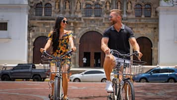 Two people on bicycles with baskets, parked in front of a historic building with arched windows and a large wooden door.