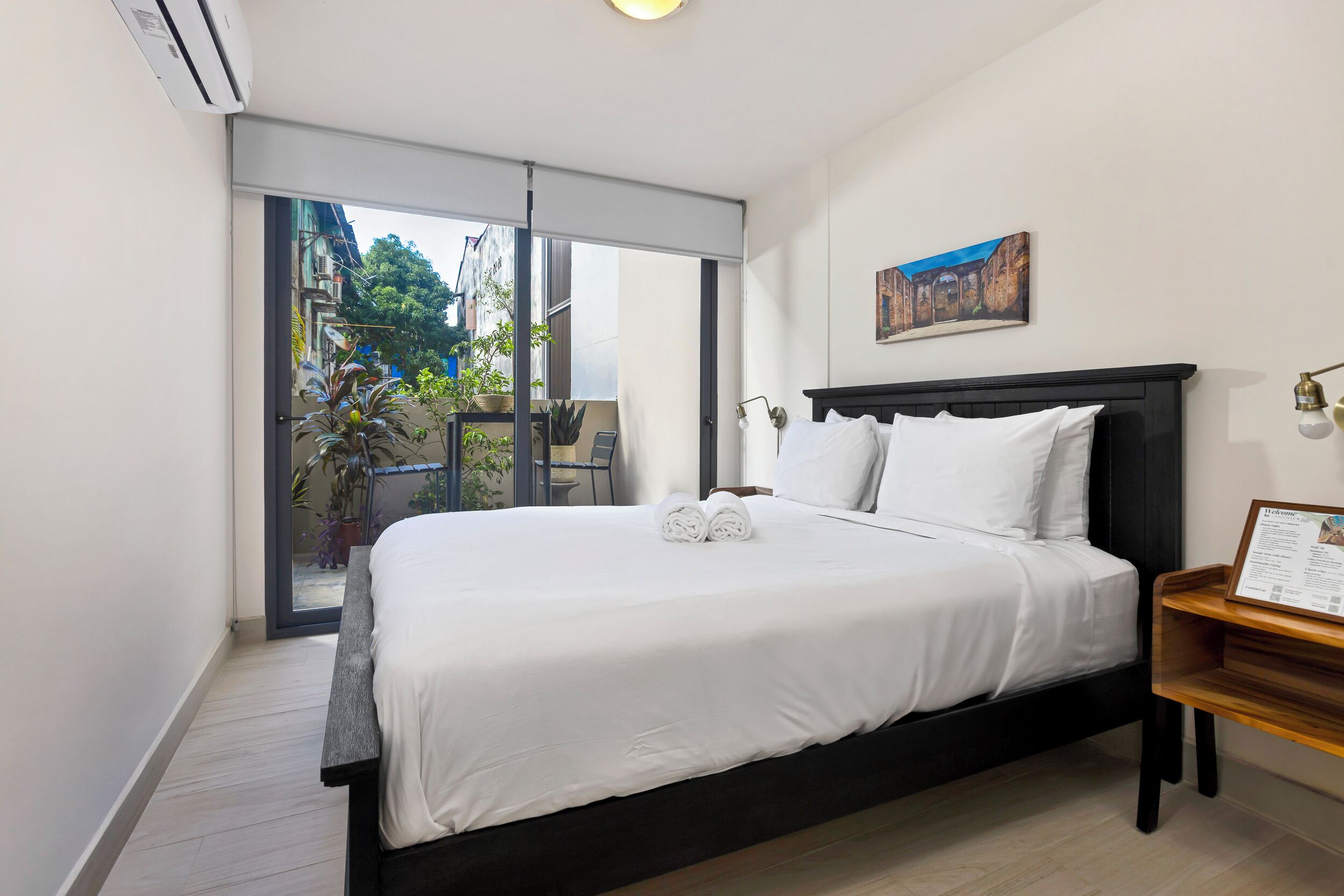 A neatly made bed with white linens, a black headboard, and a small wooden nightstand with a framed item.