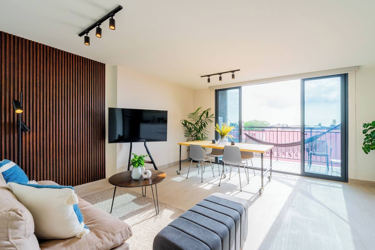 A modern living room with a flat-screen TV, a wooden accent wall, a dining area with a table and chairs, and a balcony with a view.