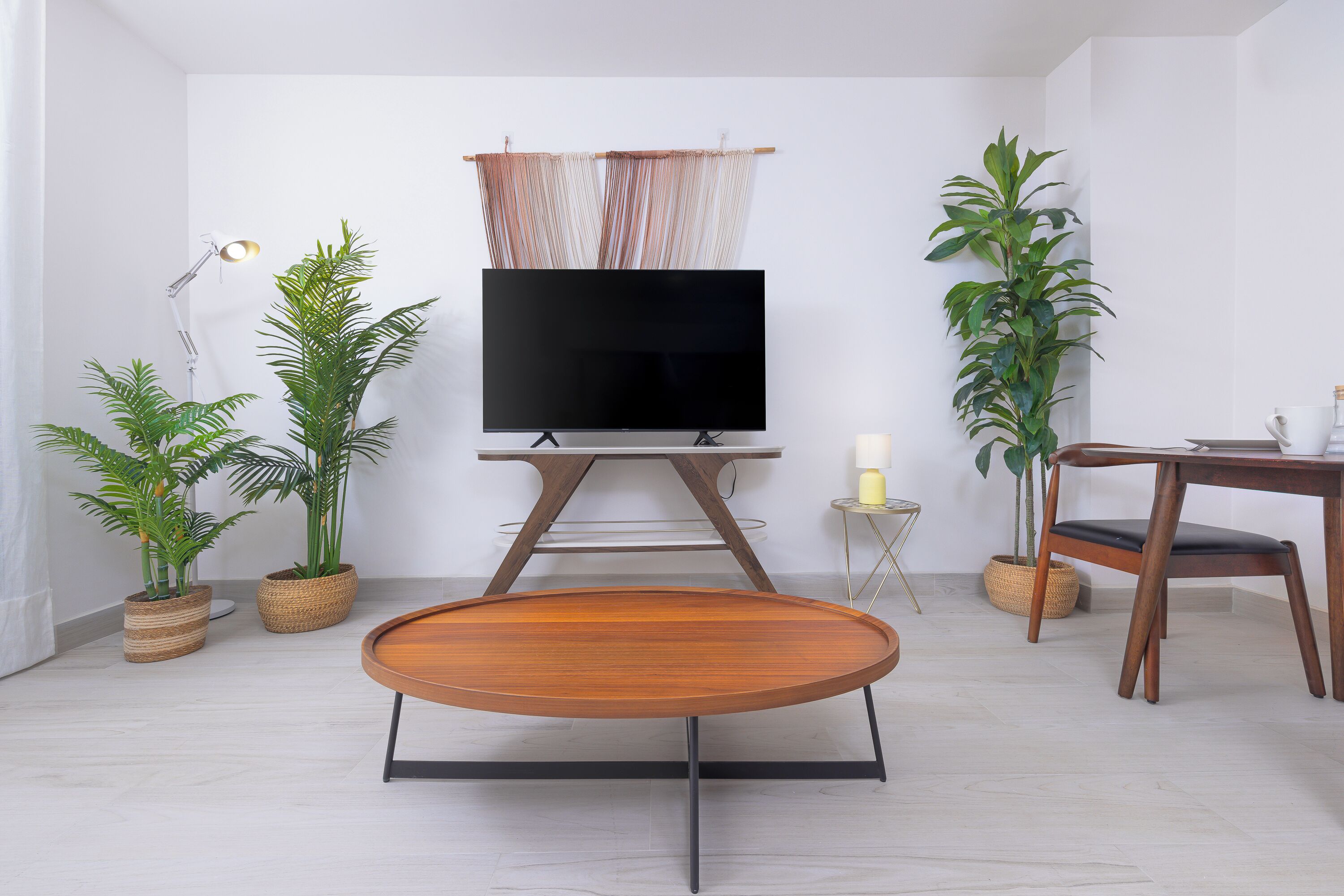 A modern living room with a flat-screen TV on a wooden stand, a round wooden coffee table, two potted plants, a small side table with a candle, and a wooden dining table with a chair.