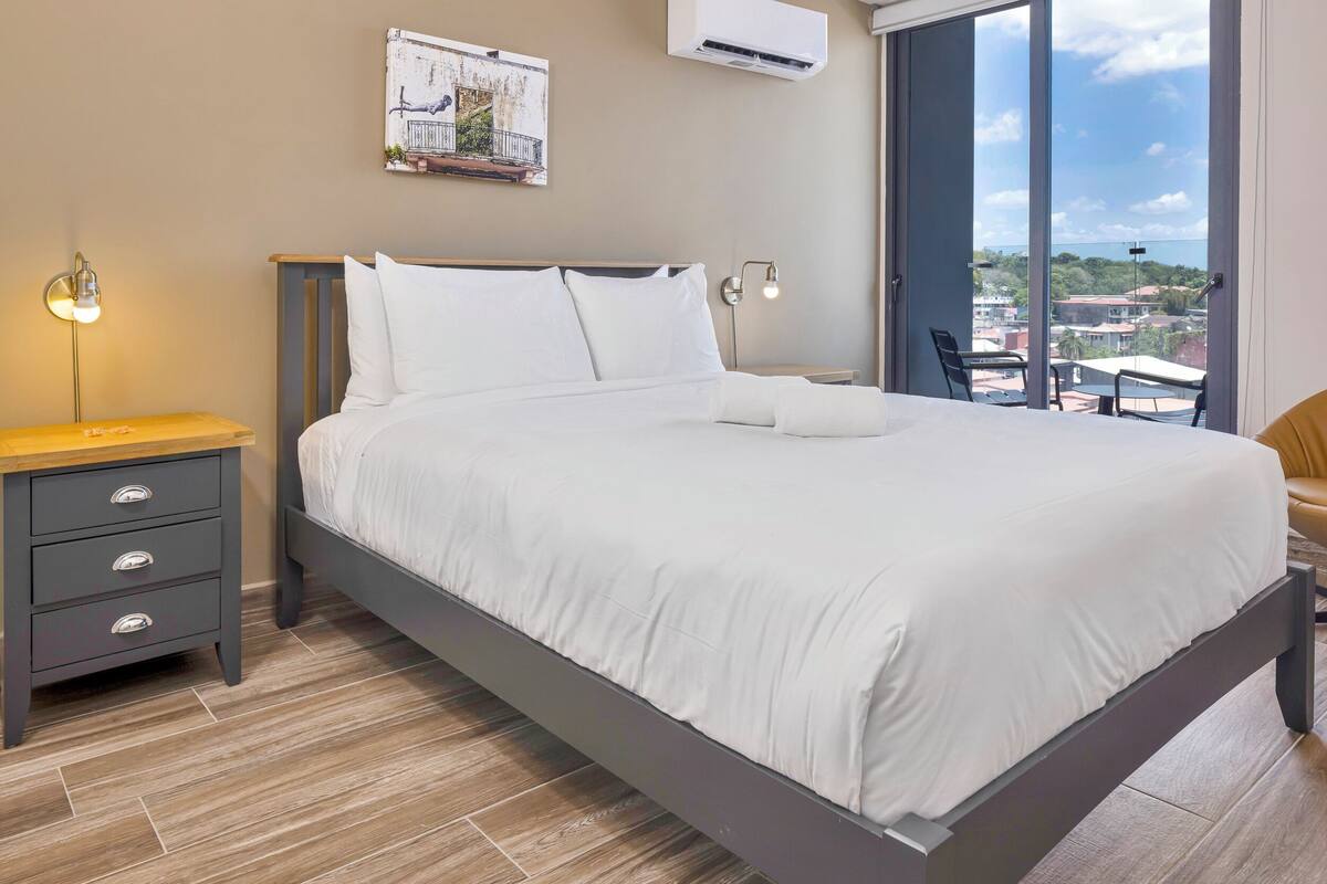 A modern bedroom with a large bed, a nightstand, a wall-mounted air conditioner, and a view of the city through large windows.