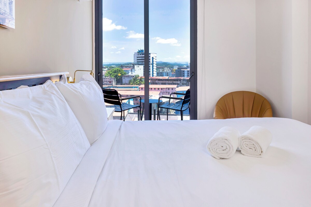 A hotel room with a large bed, two chairs, and a view of the cityscape through large windows.