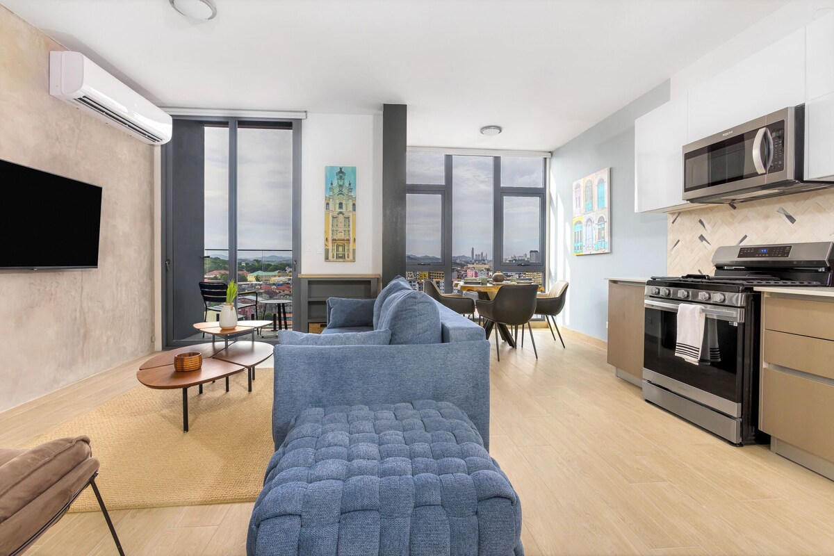 A modern living room with a large sofa, a dining area, and a kitchen with a stove and microwave.
