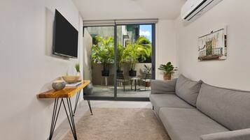 A modern living room with a grey sofa, a wooden side table with decorative items, a flat-screen TV mounted on the wall, and a large sliding glass door leading to an outdoor area with plants and furniture.