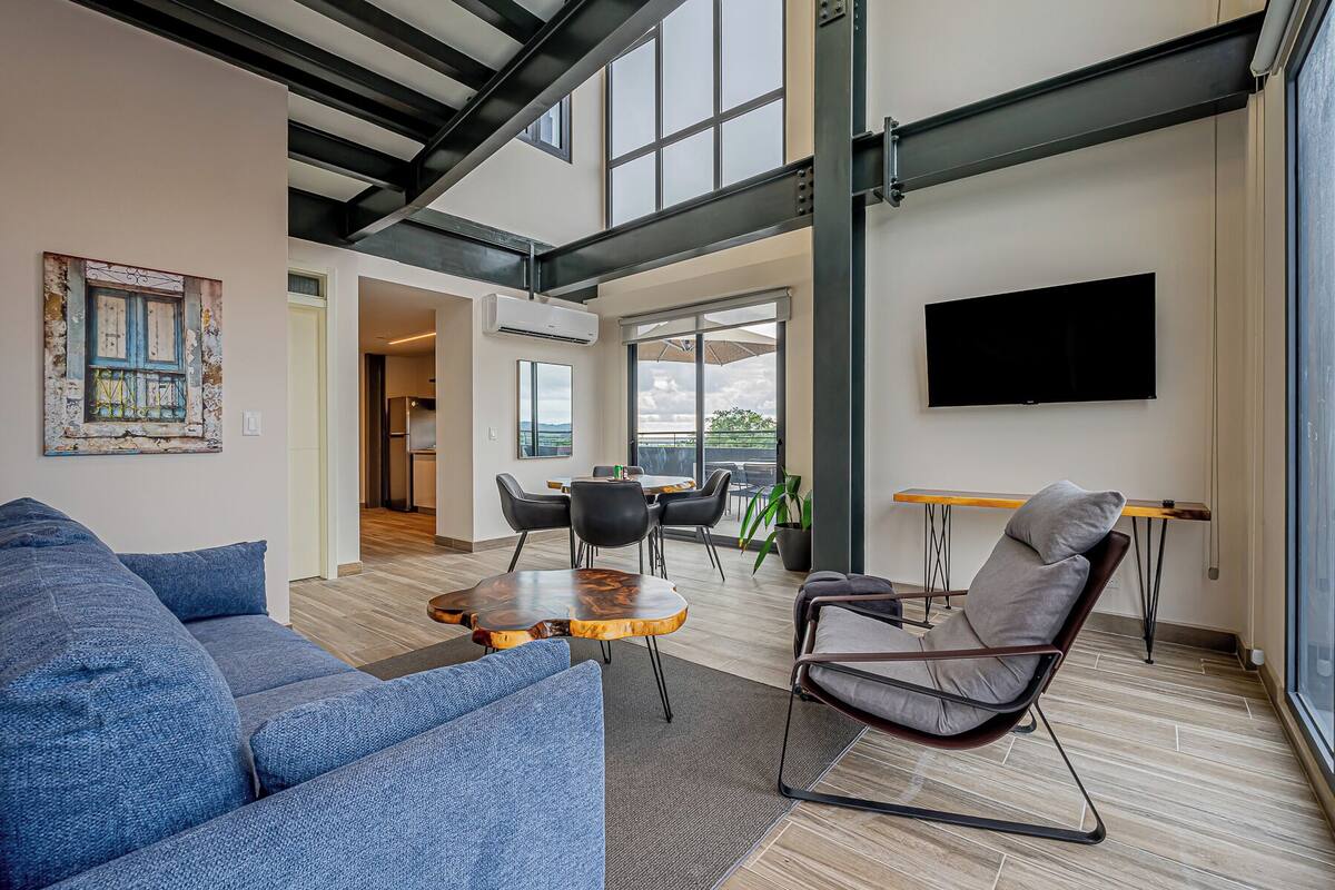 A modern living room with a large sofa, a coffee table, and a chair. There is a dining area with a table and chairs, and a TV mounted on the wall.