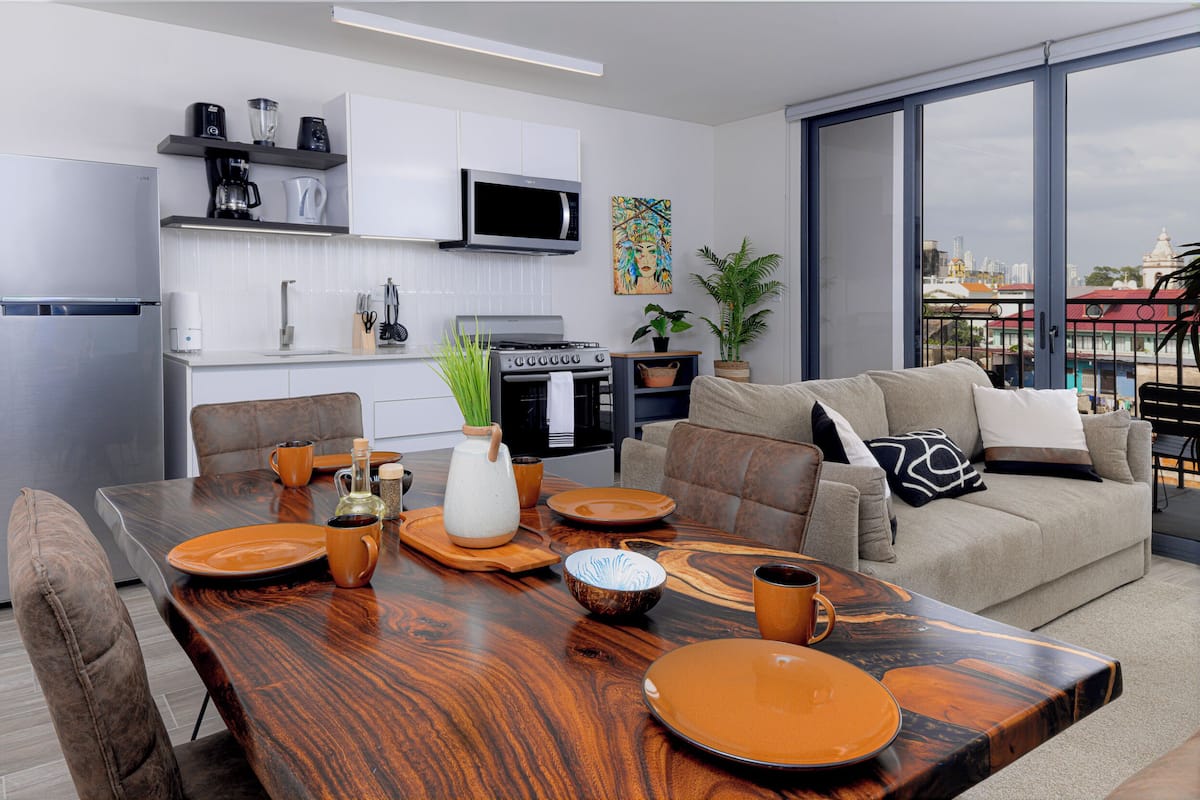 A modern kitchen with white cabinets, a microwave, and a stove. There is a dining area with a wooden table set for four, and a sofa with cushions.