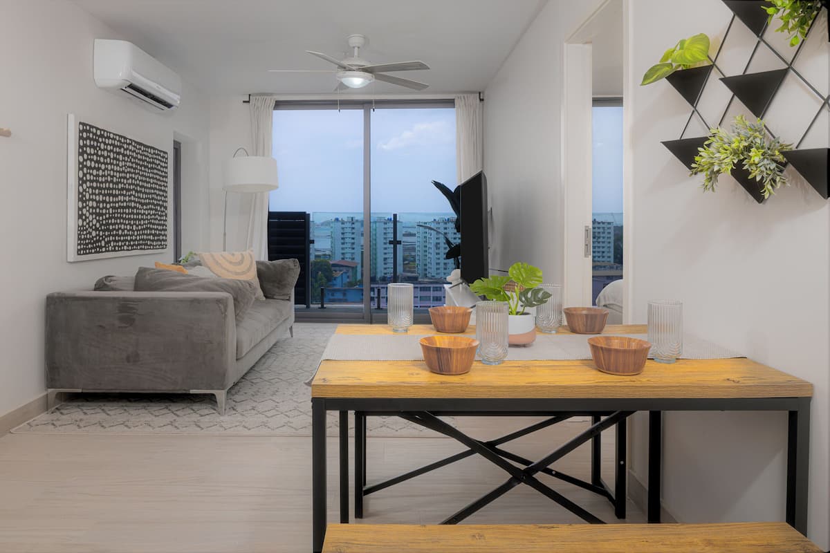 A modern living room with a grey sofa, a wooden dining table, and a view of the city through large windows.