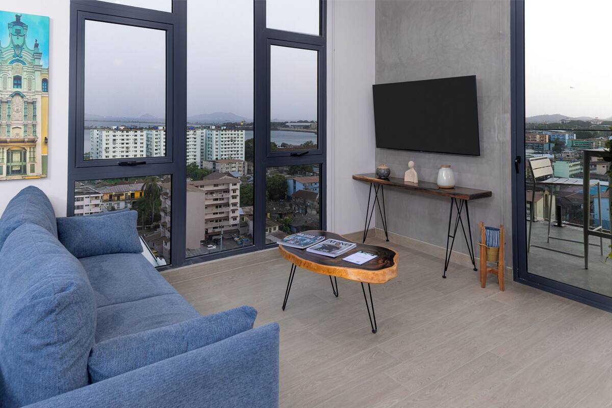 A modern living room with a large sofa, a wooden coffee table with magazines, a flat-screen TV, and a balcony with a view of the city.