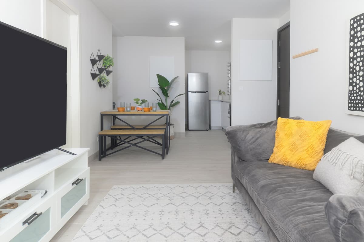 A modern living room with a flat-screen TV, a grey sofa with yellow and white cushions, a dining area with a wooden table and chairs, and a refrigerator in the background.