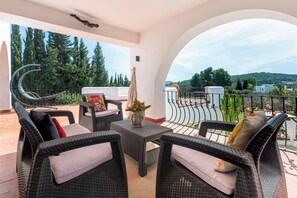 A balcony with wicker furniture, a decorative vase, and a view of trees and hills.