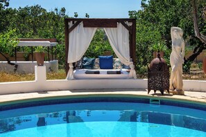 Een ontspanningsgebied bij het zwembad met een bed, een beeld en een pergola met gordijnen.