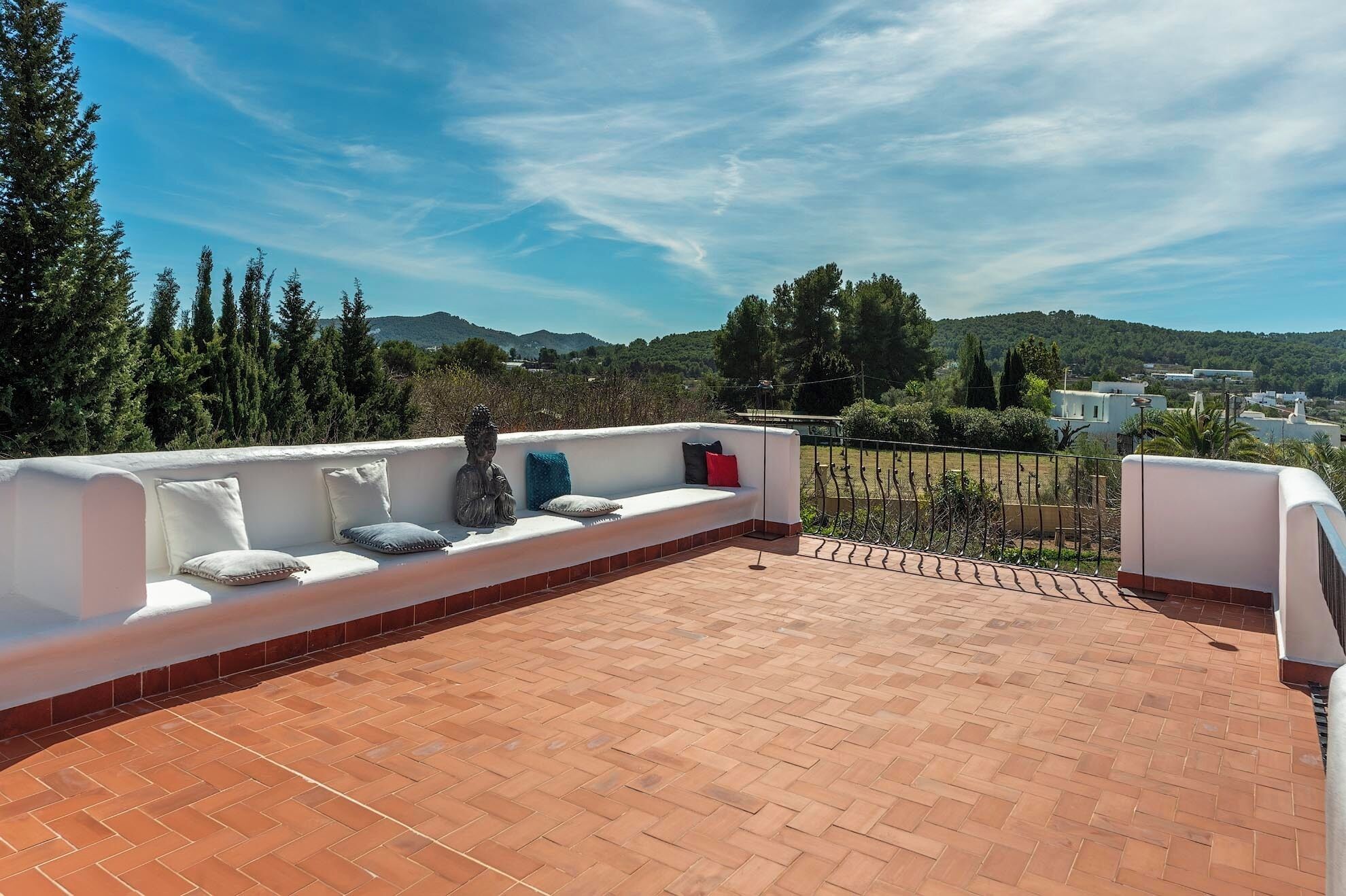 A rooftop terrace with a white sofa and cushions, a statue, and a view of a green landscape with trees and buildings in the distance.