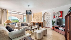 A spacious living room with a sofa set, a coffee table, ottomans, a fireplace, a TV, and a large window offering a view of greenery. - Villa Can Truyet - Four Bedroom Villa, Sleeps 7 (Santa Eulària des Riu)